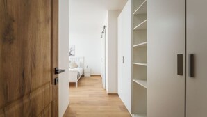 Ein Flur mit einer Holztür, einem weißen Wandschrank mit Regalen, einem Bett mit weißem Kopfteil und einem Nachttisch.