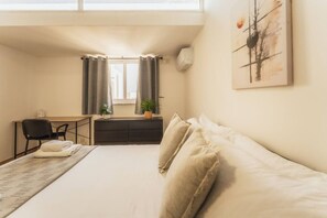 A hotel room with a bed, a desk, a chair, a small table, a plant, and a painting on the wall. - Gozo Penthouse with Terrace, BBQ & Private Roof (Gozo)