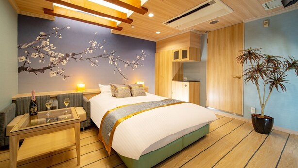 A modern hotel room with a large bed, a bedside table with a bottle and glass, a wall-mounted air conditioner, and a potted plant. - Noblesse (Kobe)