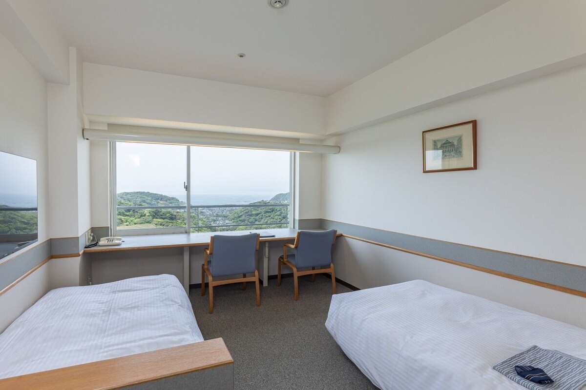 Ein Hotelzimmer mit zwei Einzelbetten, einem Schreibtisch mit Stuhl, einem Fernseher und einem großen Fenster mit Blick auf die Landschaft.