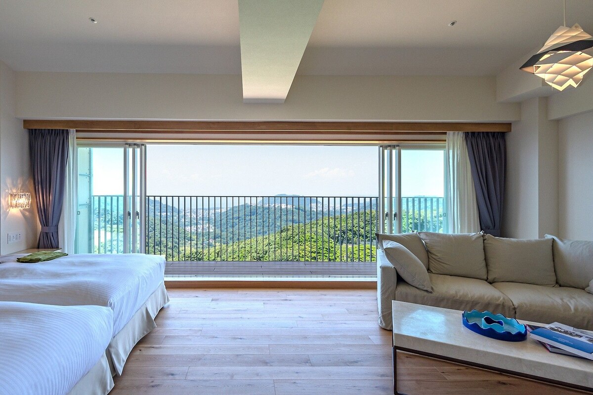 Habitación de hotel con dos camas, un sofá, una mesa de centro y un balcón con vistas al paisaje.