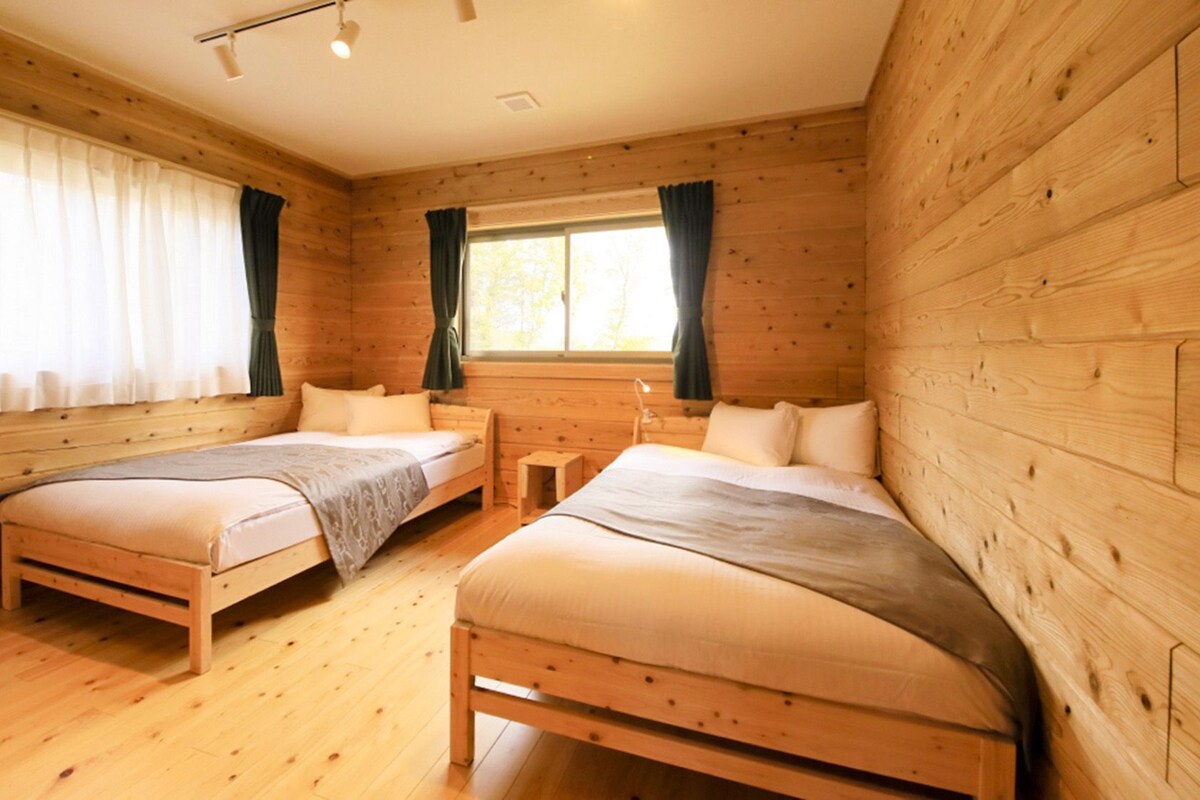 A wooden cabin room with two beds, a small table, and large windows with curtains.