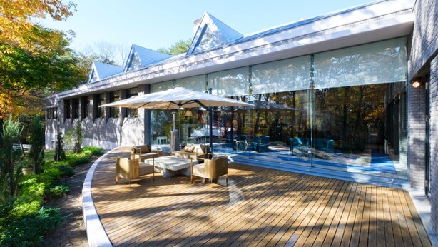 Un patio moderno al aire libre con piso de madera, asientos exteriores y grandes ventanales.