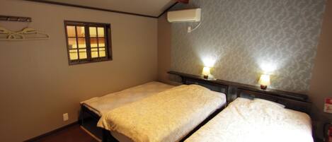 A room with two single beds, wooden beams on the ceiling, a window, and an air conditioning unit.