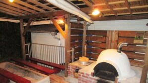 A rustic outdoor kitchen with a brick oven, wooden benches, and a hanging light fixture. - Villa Ozawa (Shimoda)