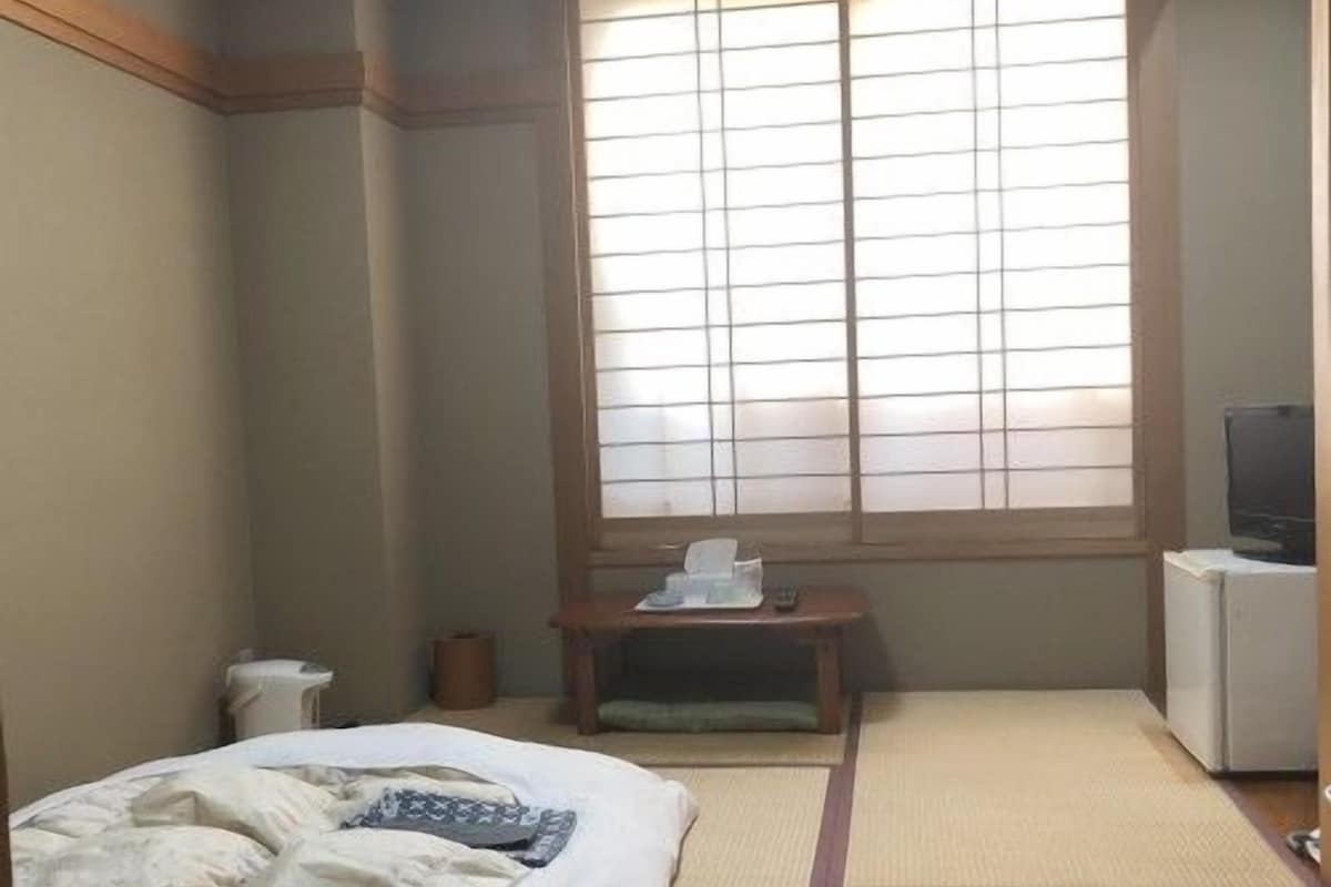 A room with a bed, a small table with a remote control, a trash bin, and a window with a view.