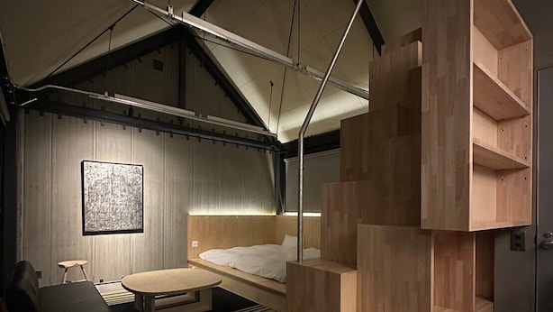 Eine moderne, mehrstöckige Holzkonstruktion in einem Raum mit schräg gedachter Decke, einem Bett, einem Tisch und einem an der Wand befestigten Kunstwerk.