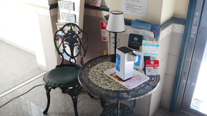 Una esquina de una habitación con una silla, una mesa con una lámpara y varios objetos sobre la mesa, incluyendo una caja y un letrero.