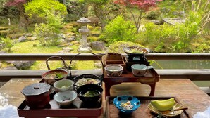 Un ambiente tradicional japonés para la ceremonia del té, con diferentes utensilios y vajilla sobre una mesa de madera, con vistas a un jardín que incluye un farol de piedra y abundante vegetación.