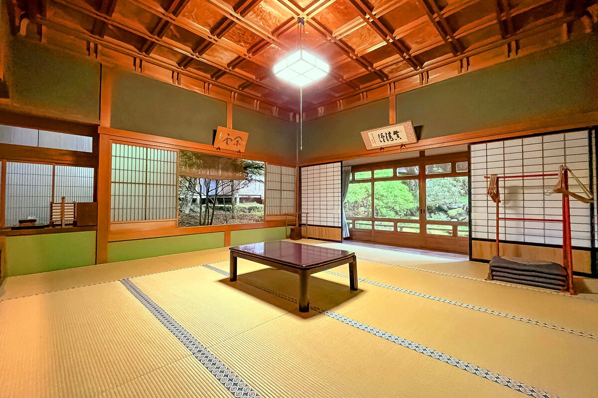 A traditional Japanese room with tatami flooring, a low wooden table, sliding shoji doors, and a view of greenery outside.