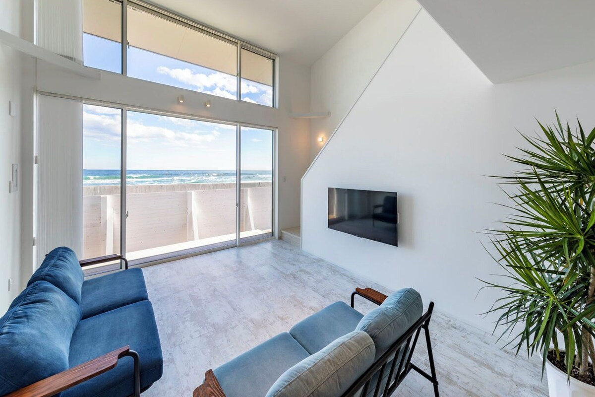 A spacious living area with a large sliding glass door leading to a balcony, a flat-screen TV mounted on the wall, and a potted plant.