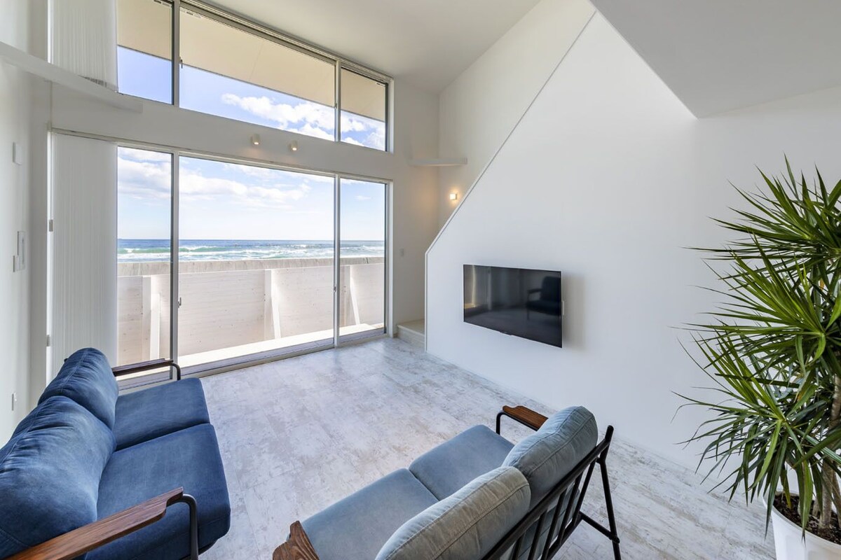 A spacious living area with a large sliding glass door leading to a balcony, a flat-screen TV mounted on the wall, and a potted plant.