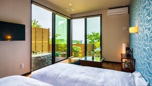 Ein modernes Hotelzimmer mit einer großen Schiebeglasdoor zum Balkon, einem Bett, einem kleinen Tisch mit einer Lampe und einem an der Wand montierten Fernseher.