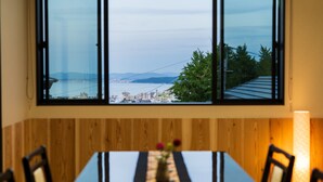 Ein Essbereich mit Tisch und Stühlen, ein Fenster mit Blick auf eine Stadtlandschaft und eine Brücke sowie eine Vase mit Blumen auf dem Tisch.
