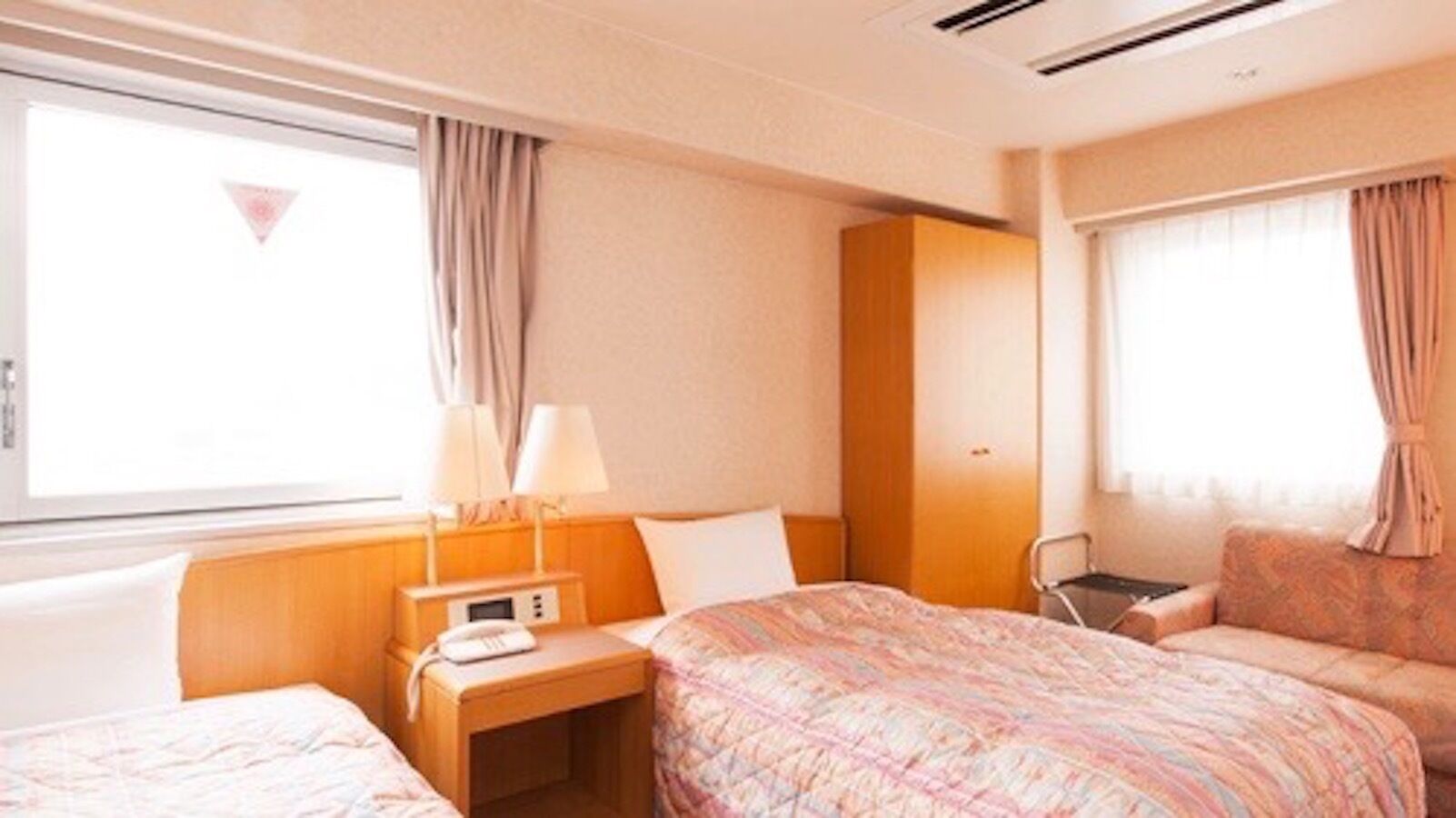 Ein Hotelzimmer mit zwei Betten, einem Nachttisch mit Lampe, einem kleinen Tisch mit Telefon und einem Fenster mit Vorhängen.