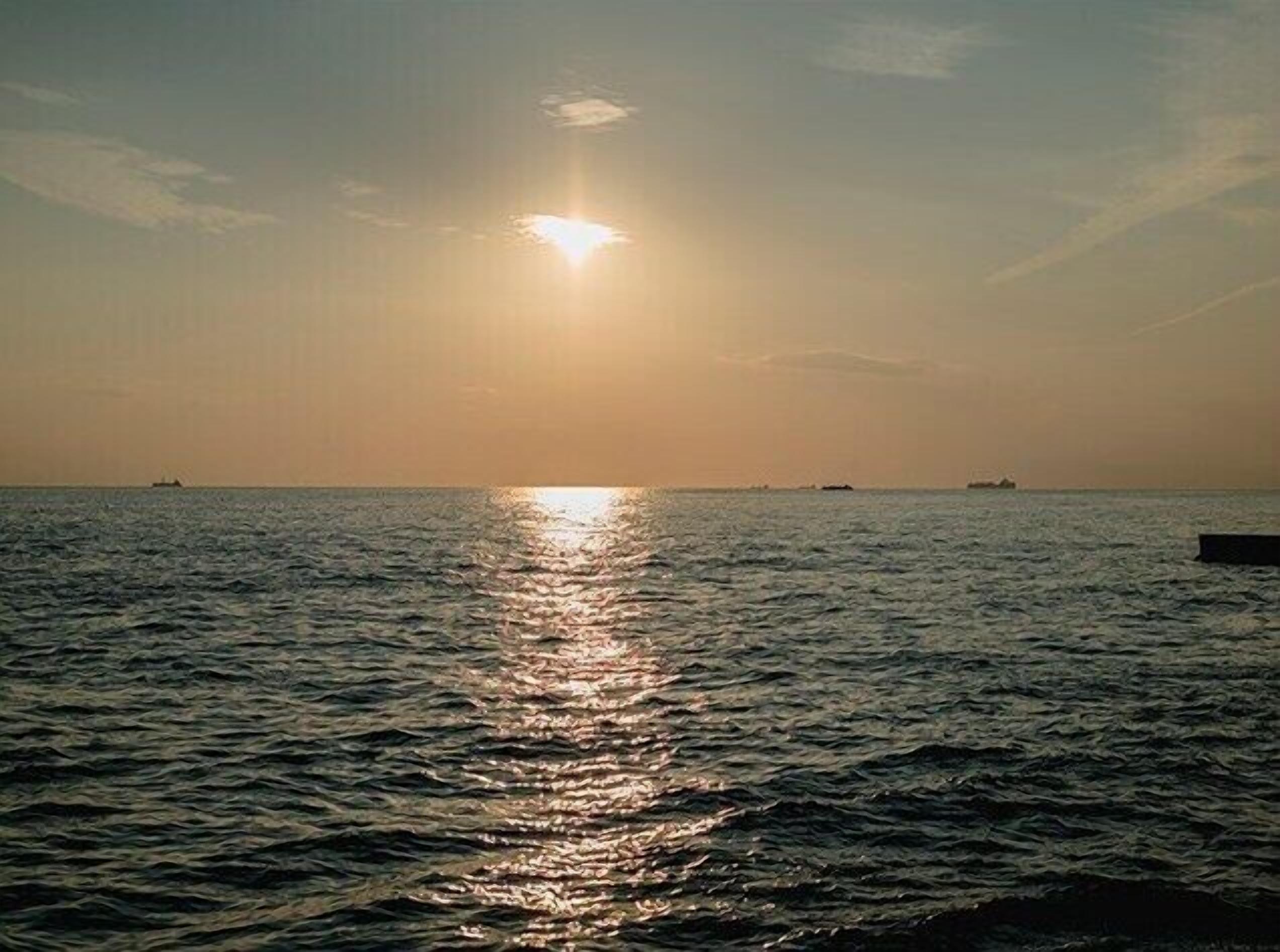 Une mer calme, le soleil se reflétant sur l’eau, des bateaux à l’horizon et un ciel dégagé.