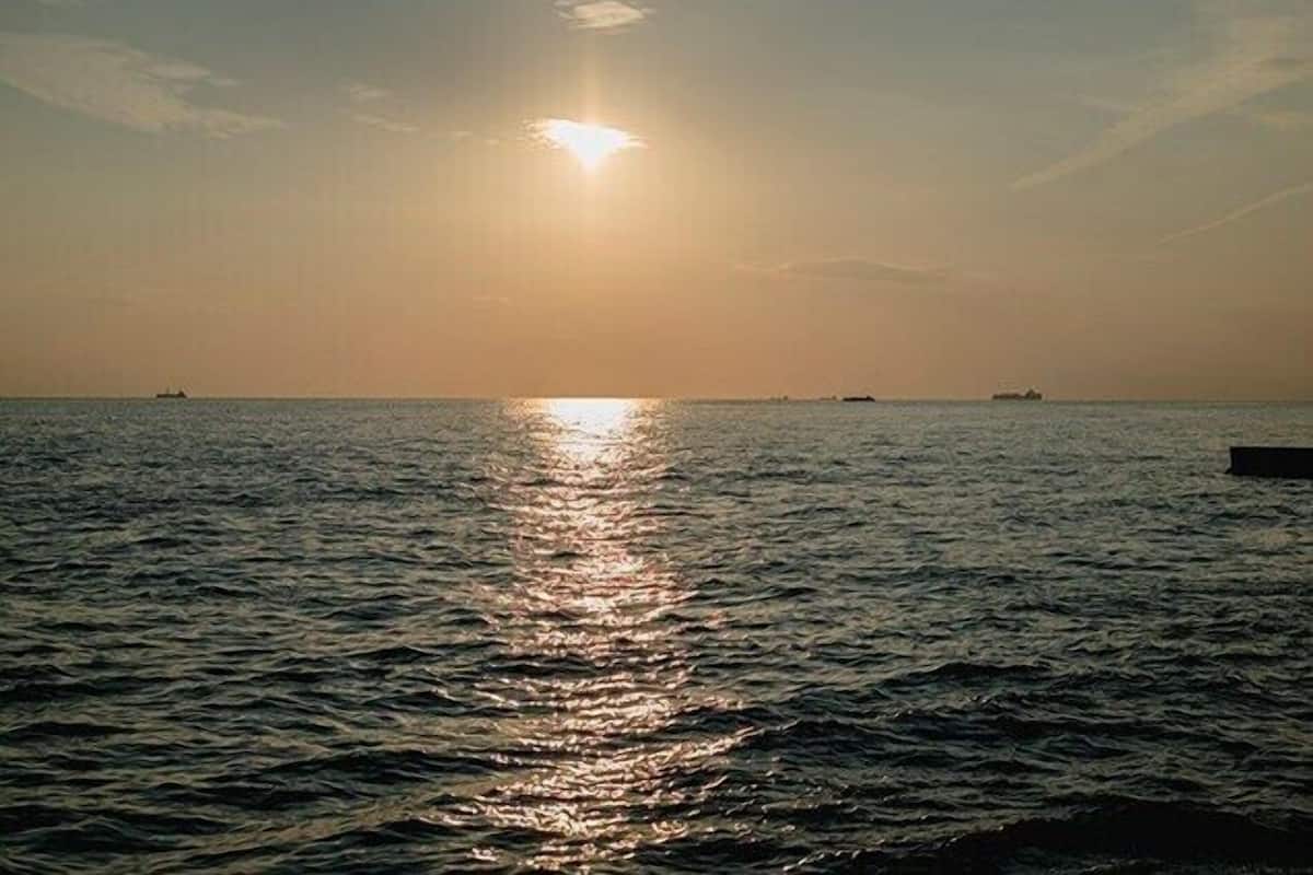 Mar tranquila con el sol reflejándose en el agua, barcos a lo lejos y un cielo despejado.