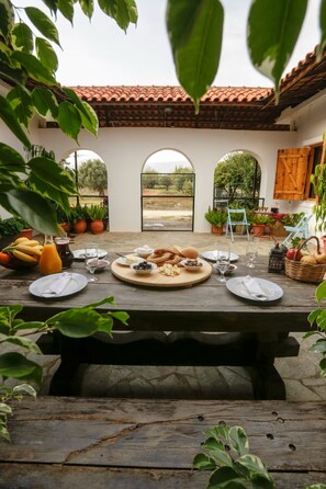 Un espacio exterior para comer con una mesa de madera rústica dispuesta para una comida, rodeado de plantas en macetas y una estructura de techo de baldosas.