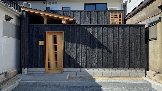 Un bâtiment moderne doté d’une façade sombre à la texture marquée, d’une porte en bois et d’une petite fenêtre.