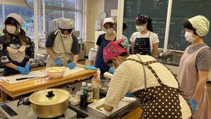 A group of people in a classroom setting, engaged in a cooking activity with a stove, pots, and ingredients on the table. - Sanriku Active (Ofunato)