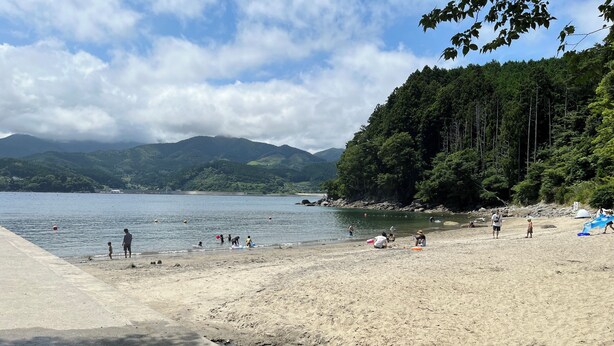A beach with a concrete walkway, a sandy shore, and a forested hillside in the background. - Sanriku Active (Ofunato)