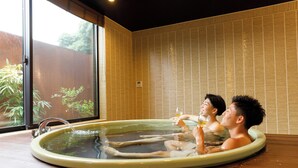 Una bañera de hidromasaje con dos personas disfrutando el agua, una ventana con persianas y vistas a la vegetación exterior.