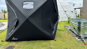Ett svart tält med ett fönster, placerat på gräset med en metallbehållare i närheten.