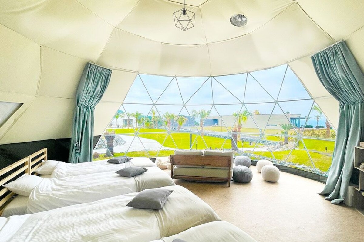 A room with three beds, a wooden headboard, a small table, and a view of a green landscape through a large window.