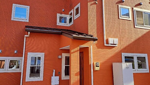 Ein Gebäude aus roten Ziegelsteinen mit mehreren Fenstern, einer sichtbaren Tür und einem kleinen Außenbereich mit einem Metallgeländer.