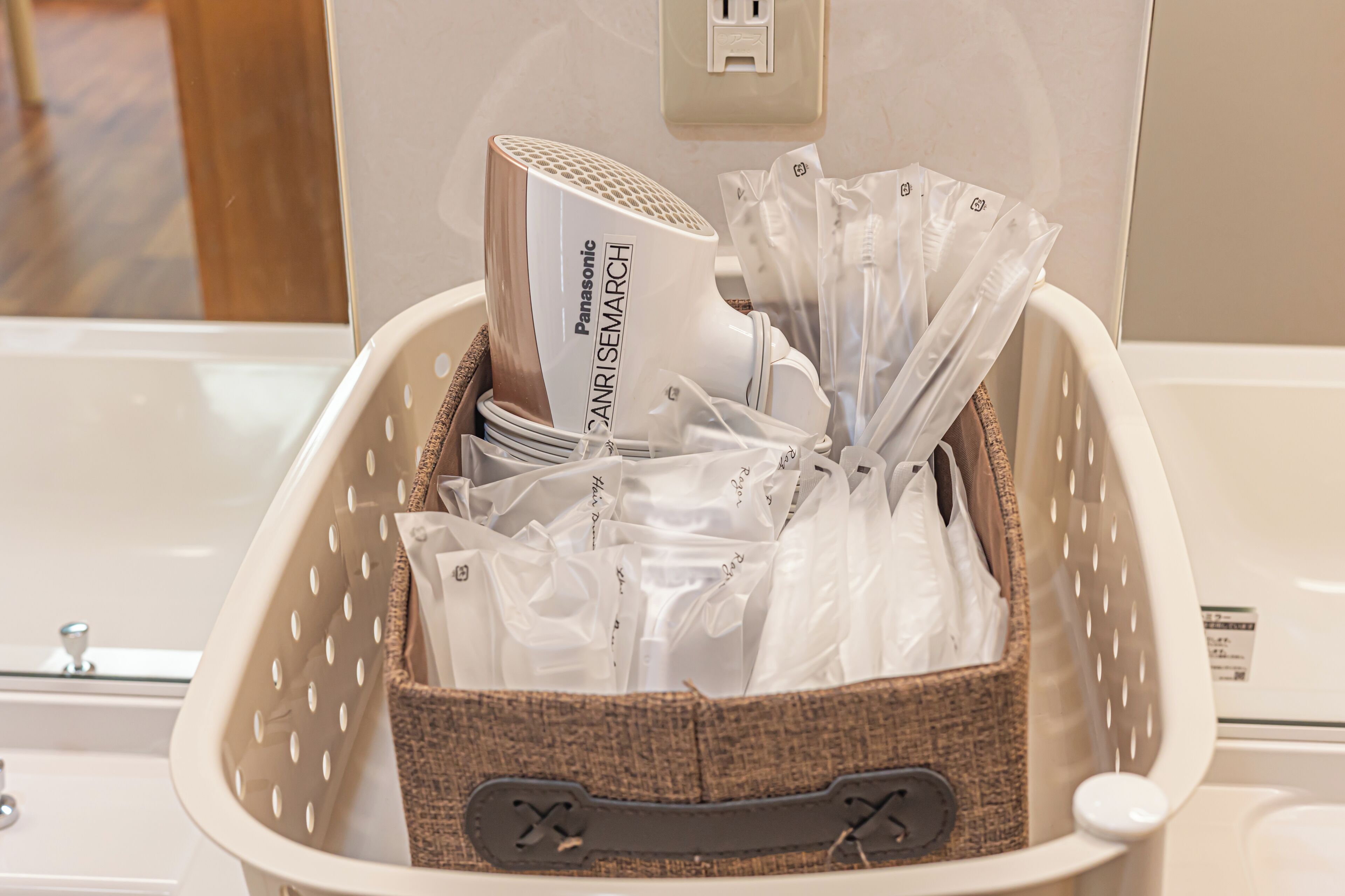 A basket containing a Panasonic hair dryer, several hair care products, and a white appliance, likely a hair straightener, in a hotel setting.
