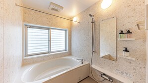 A modern bathroom with a freestanding bathtub, a shower, a window with blinds, a mirror, and a shelf with toiletries. - Sunrise Vista Nanjo (Nanjo)