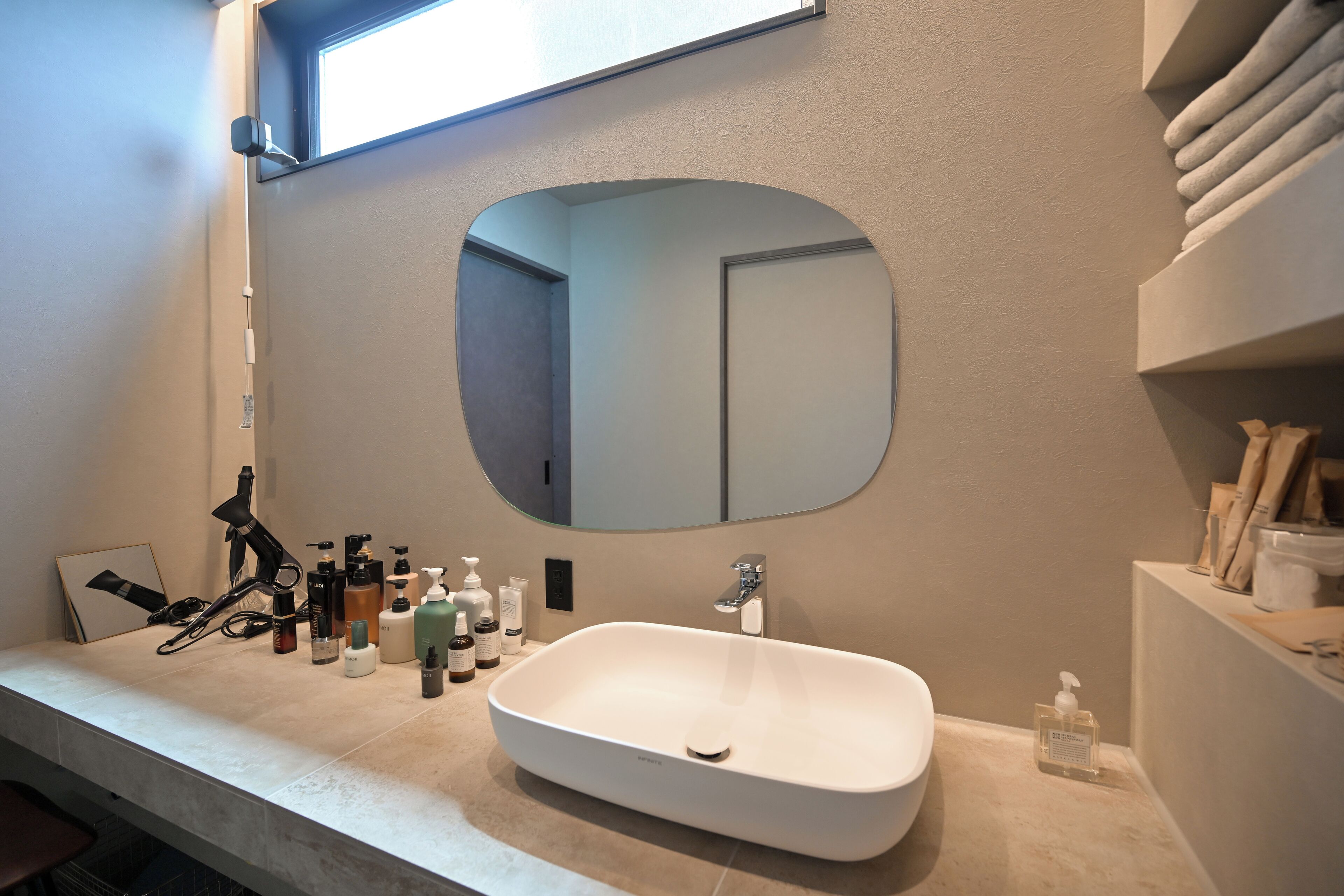 A modern bathroom with a large oval mirror, a white sink, and various toiletries on the countertop.