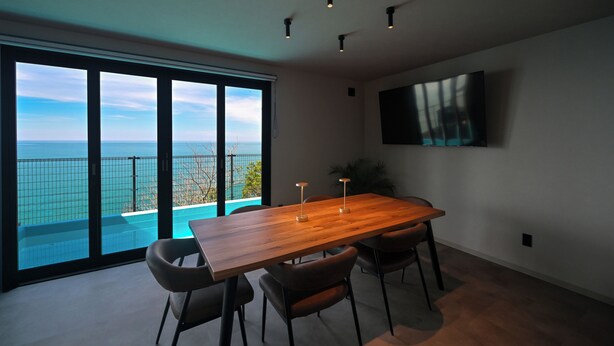 A modern dining area with a wooden table and chairs, large windows with a view of the sea, and a mounted TV on the wall. - Cliff Villa (Konan)