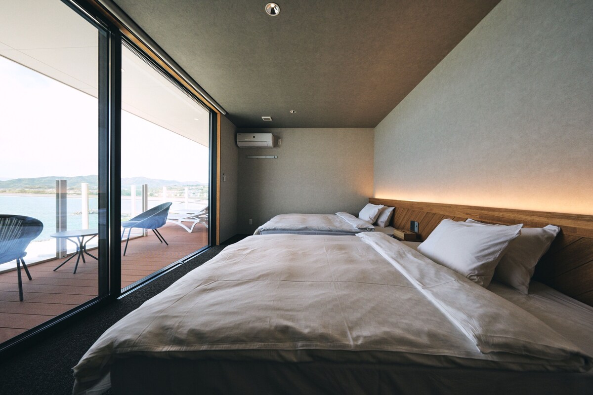Een moderne hotelkamer met een groot bed, een kleine tafel en een stoel nabij een schuifdeur die leidt naar een balkon met uitzicht op het water.