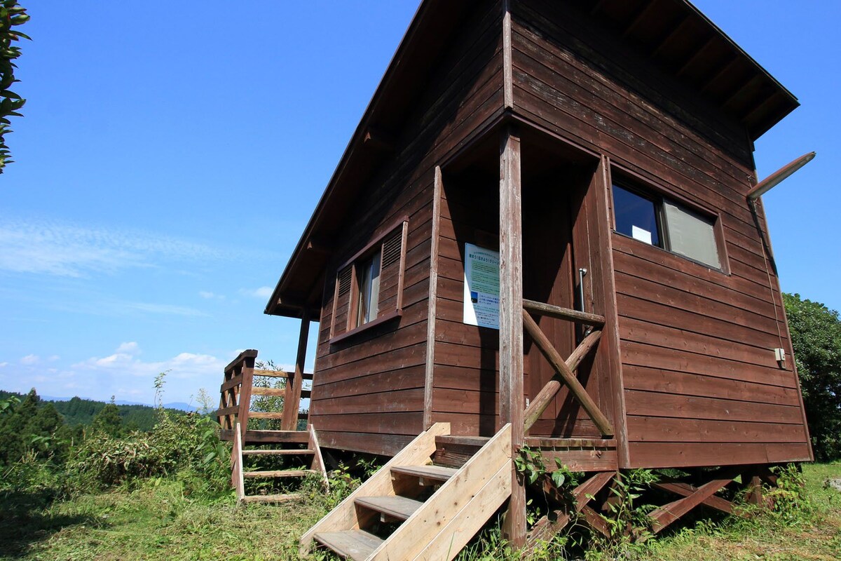 A wooden cabin with a staircase leading up to the entrance, surrounded by greenery.