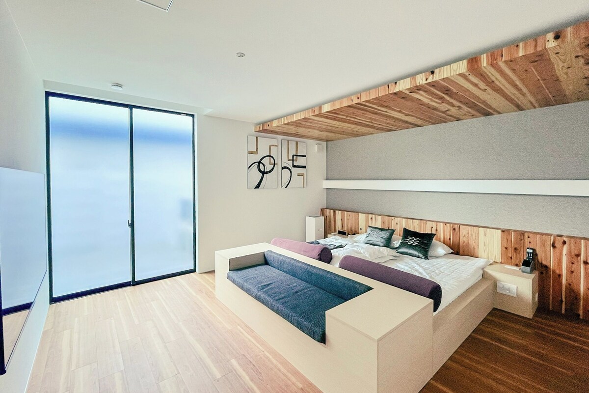 Un dormitorio moderno con una cama grande, un banco empotrado y un ventanal corredera que da a un balcón.