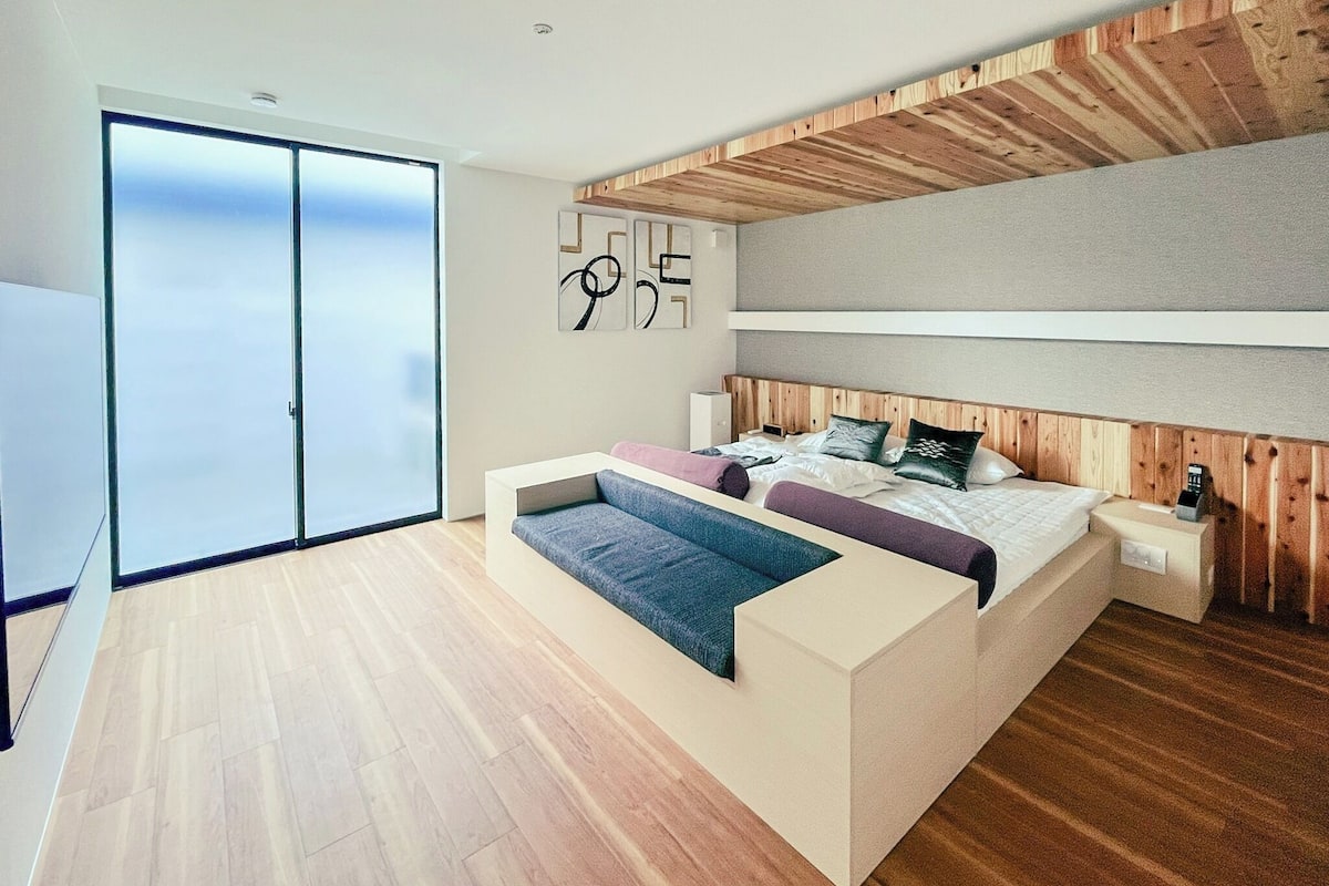 Un dormitorio moderno con una cama grande, un banco empotrado y un ventanal corredera que da a un balcón.