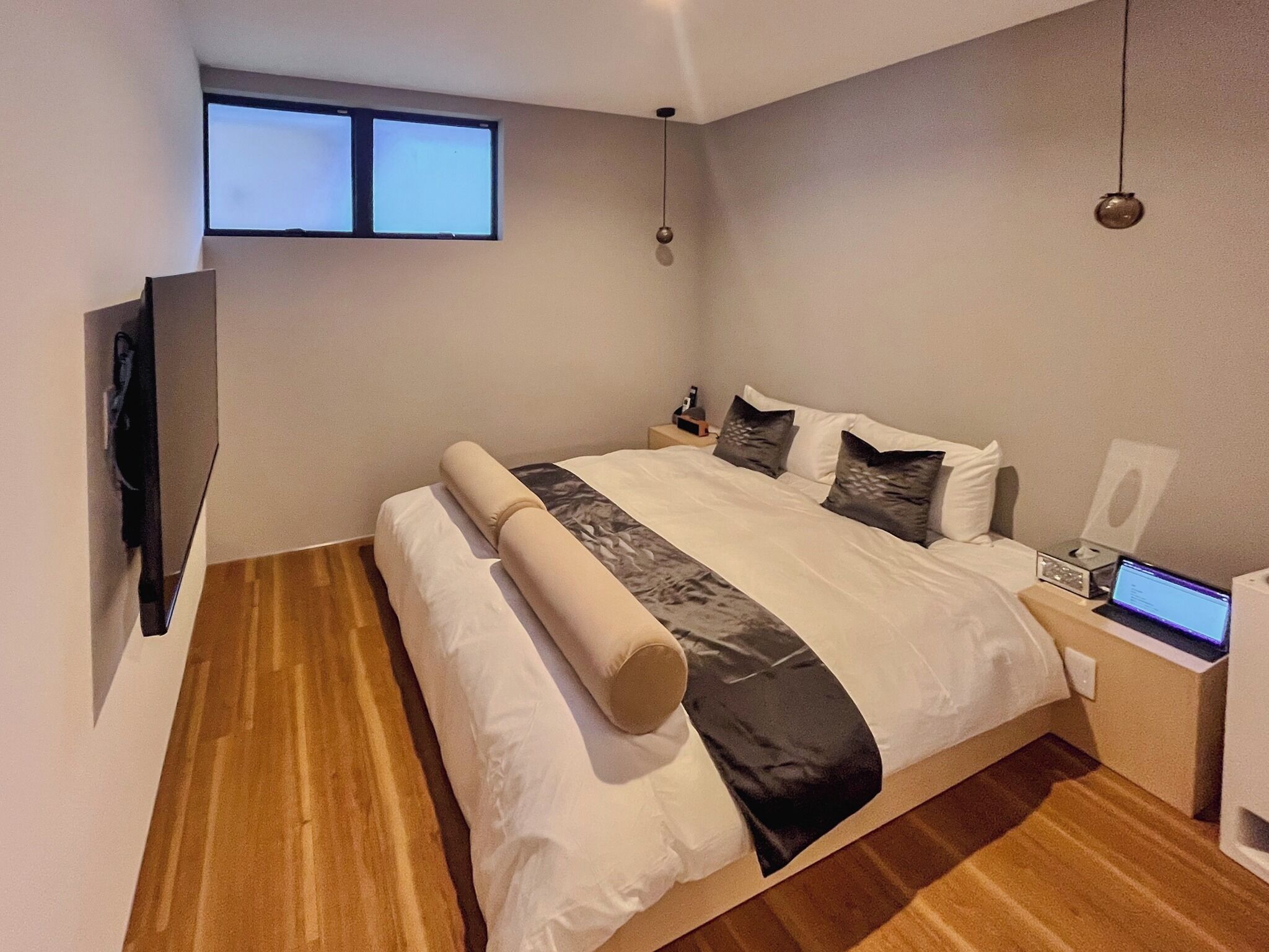 A modern bedroom with a large bed, a flat-screen TV mounted on the wall, a laptop on the bedside table, and a small table lamp.