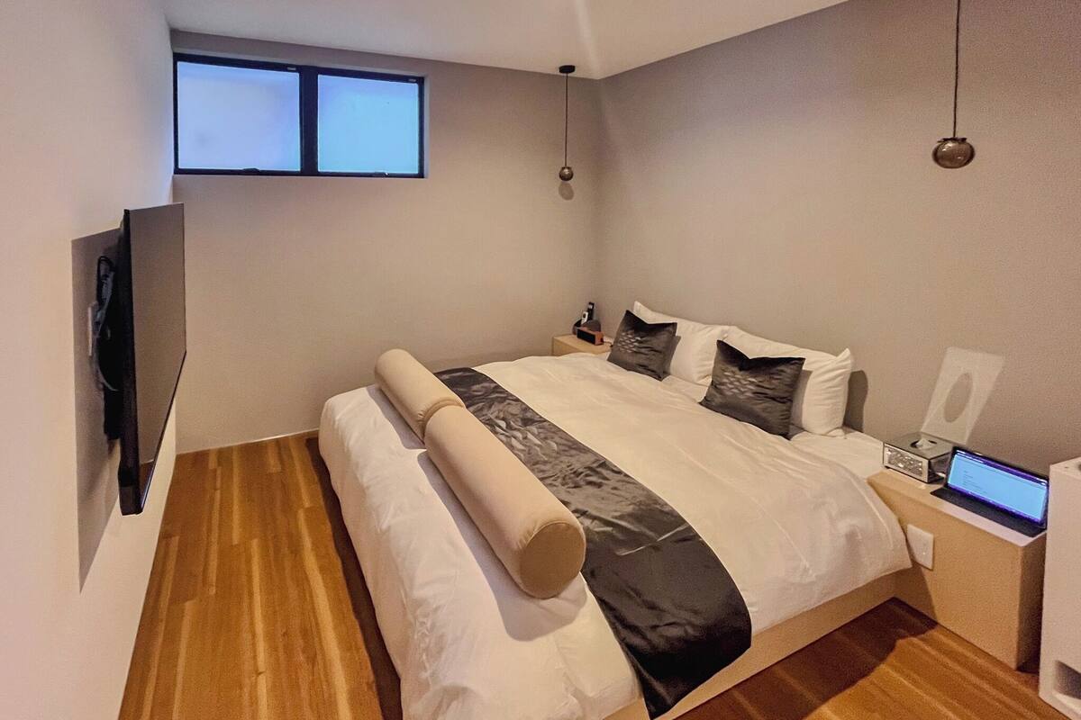 Un dormitorio moderno con una cama grande, un televisor de pantalla plana en la pared, un ordenador portátil sobre la mesita de noche y una lámpara de mesa pequeña.