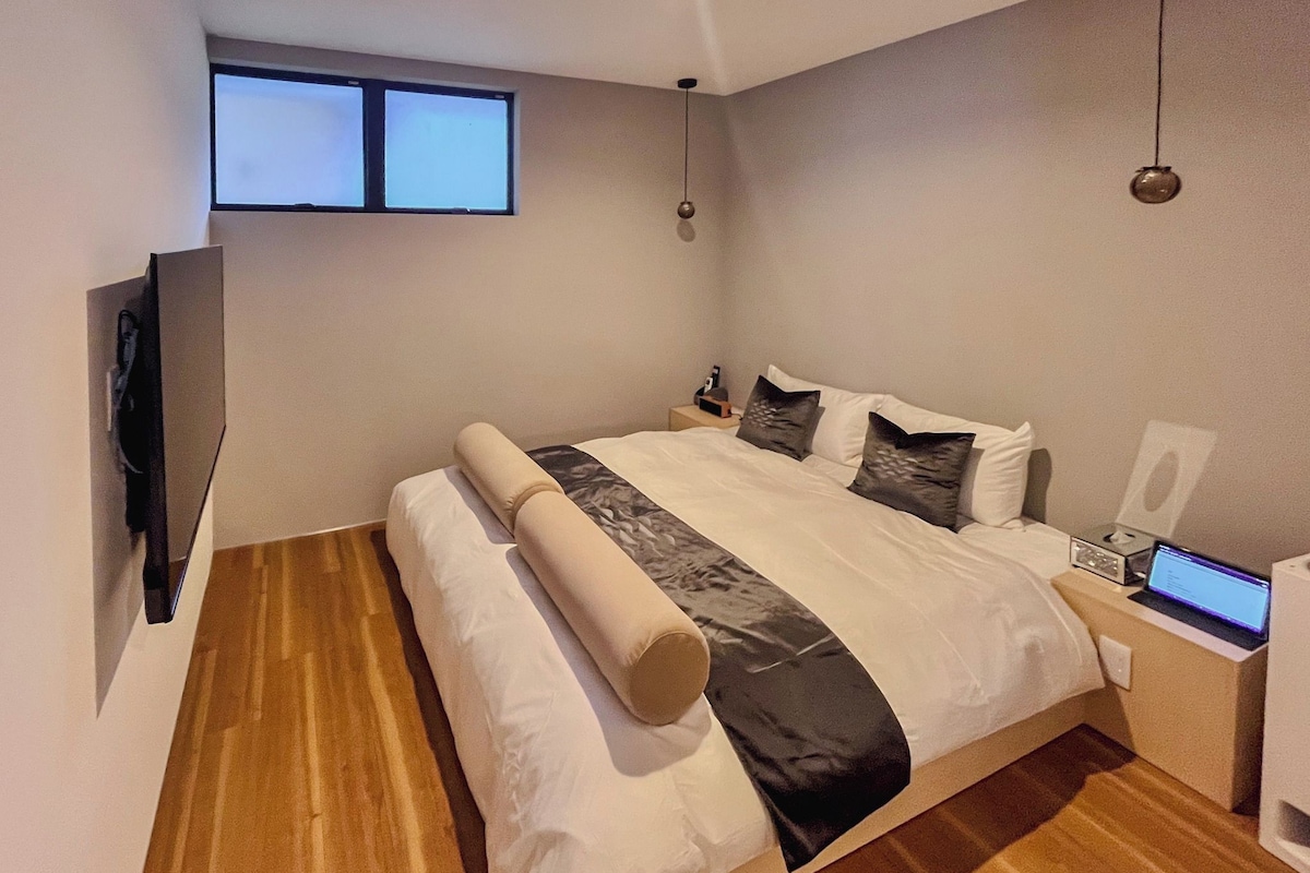 Un dormitorio moderno con una cama grande, un televisor de pantalla plana en la pared, un ordenador portátil sobre la mesita de noche y una lámpara de mesa pequeña.
