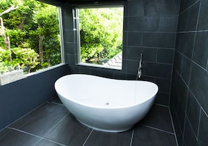 A modern bathroom with a freestanding bathtub, dark tiled walls, and a window overlooking greenery.