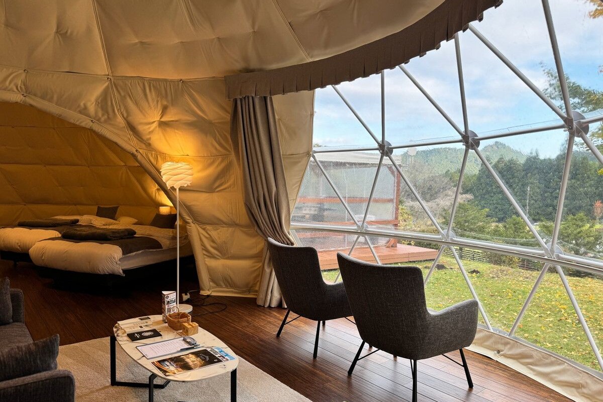 Un intérieur chaleureux avec un lit, une table sur laquelle sont posés des documents à lire et deux chaises. Une grande fenêtre offre une vue sur l’extérieur.