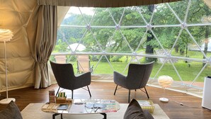 Ein Zimmer mit großem Fenster, zwei Stühlen, einem Tisch mit diversen Gegenständen und Blick auf einen Garten.