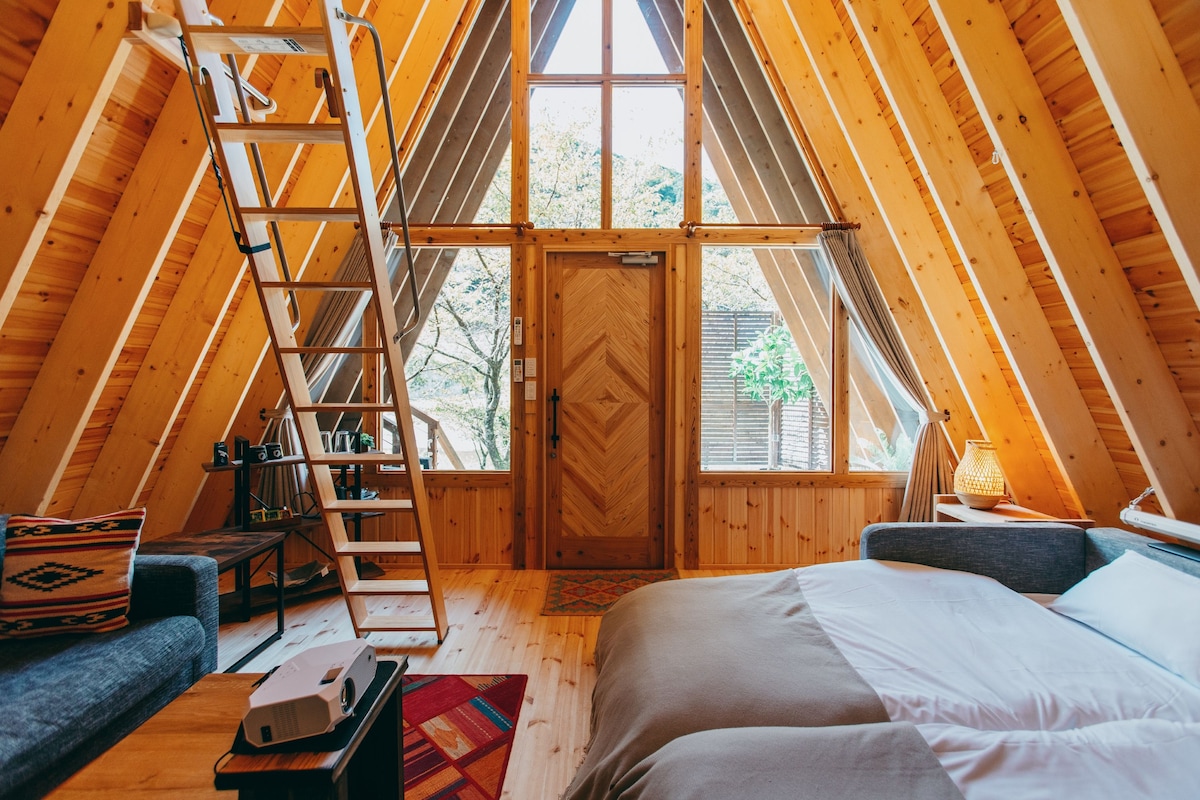 Een knus zolderkamertje met een houten ladder, een bed, een bank en een raam met uitzicht op groen.
