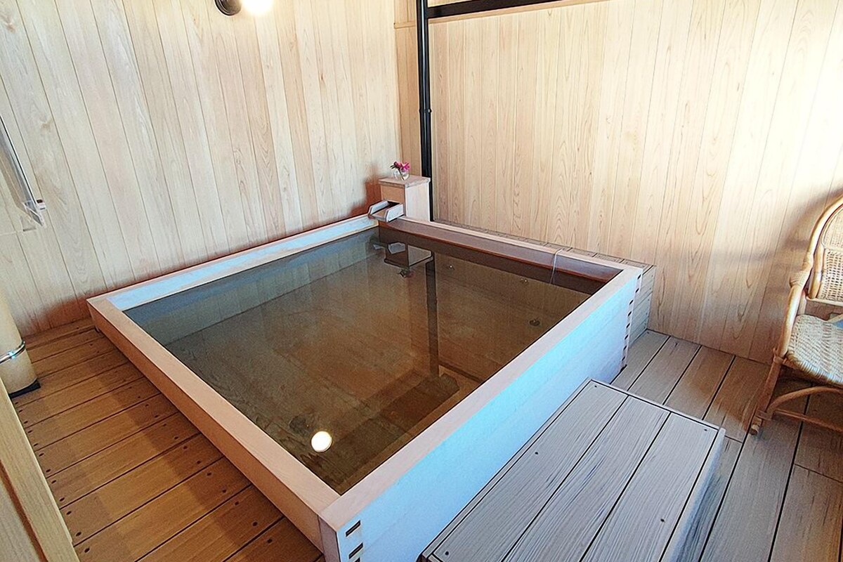 Un bain à remous en bois, entouré de parois en verre, une petite table avec un vase et un fauteuil en osier.
