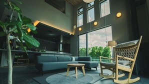 Un salon moderne avec un canapé, un fauteuil à bascule en bois et un coin repas, la cuisine étant visible au fond.