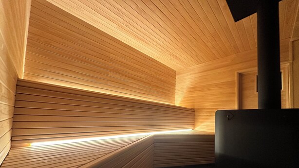 A sauna with wooden slats and a bench, a black heater, and a wooden door. - Akiya Cornerstone (Awaji)