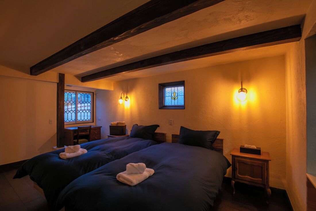 A room with two beds, wooden beams on the ceiling, a window with a stained glass design, and wall-mounted lamps.