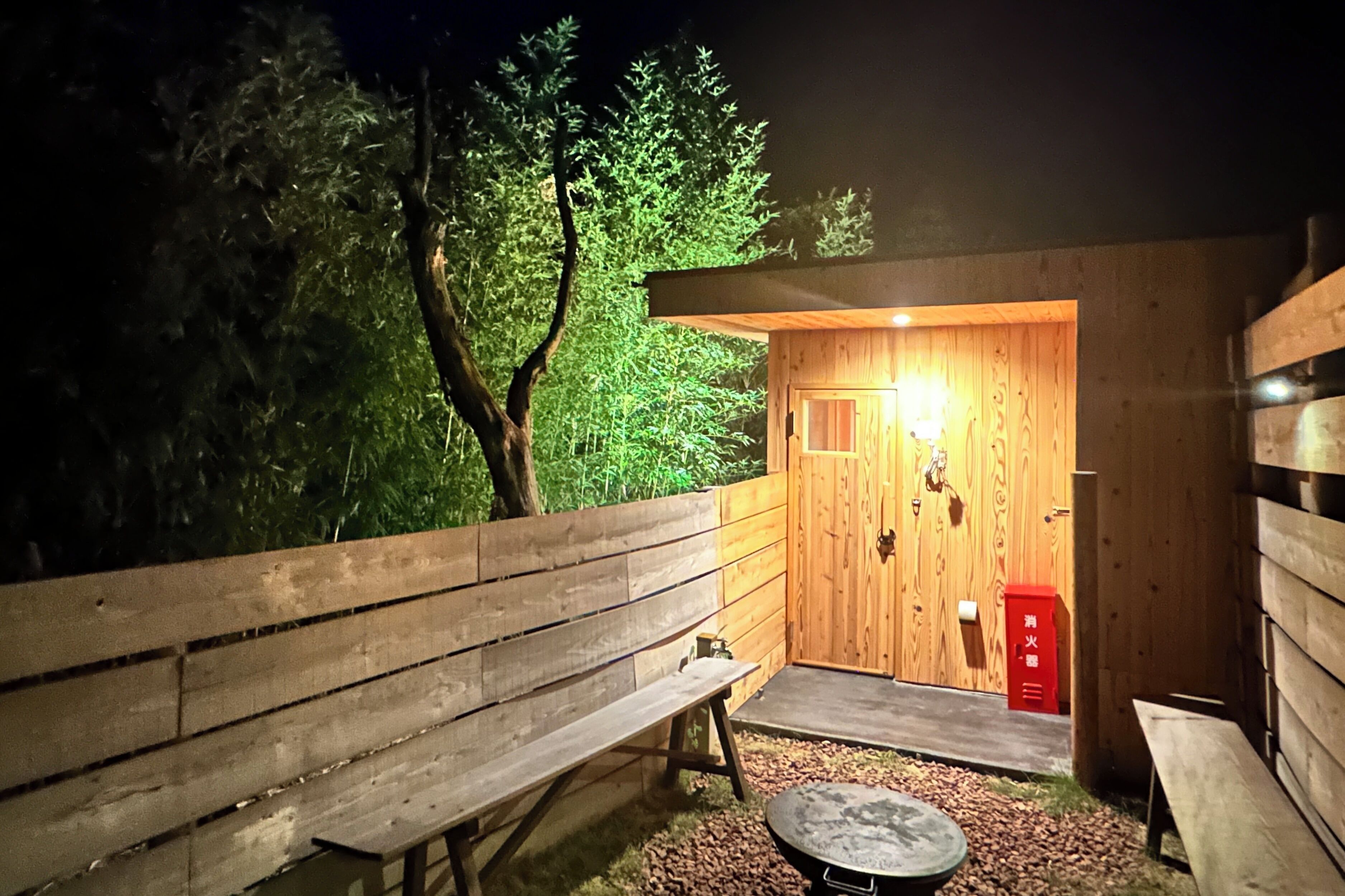 A wooden cabin with a bench and a table outside, surrounded by trees.