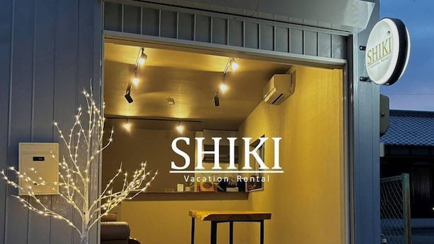 Ein Hotel Eingangsbereich mit einer Rezeption, einem Schild mit der Aufschrift "SHIKI Vacation Rental" und einem kleinen Baum mit Lichterketten.